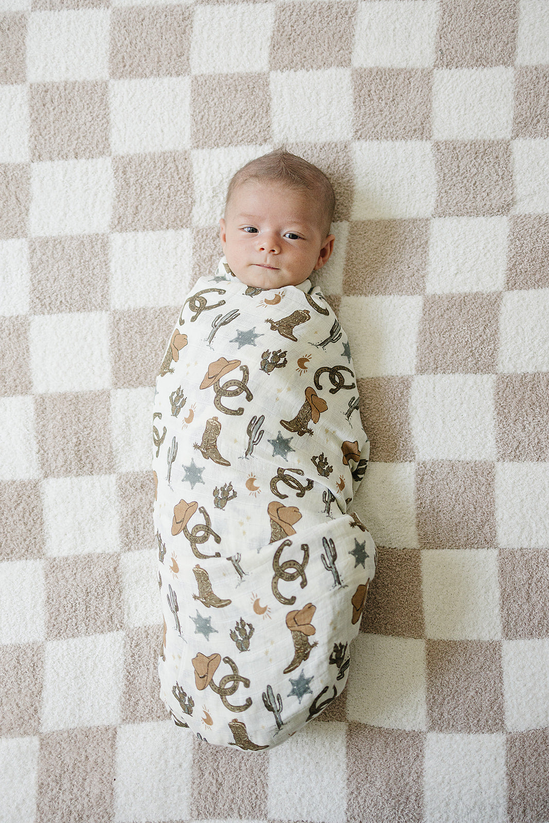 Baby wrapped in Outlaw Cowboy Muslin Swaddle Blanket, featuring cowboy-themed designs, on a checkered blanket.