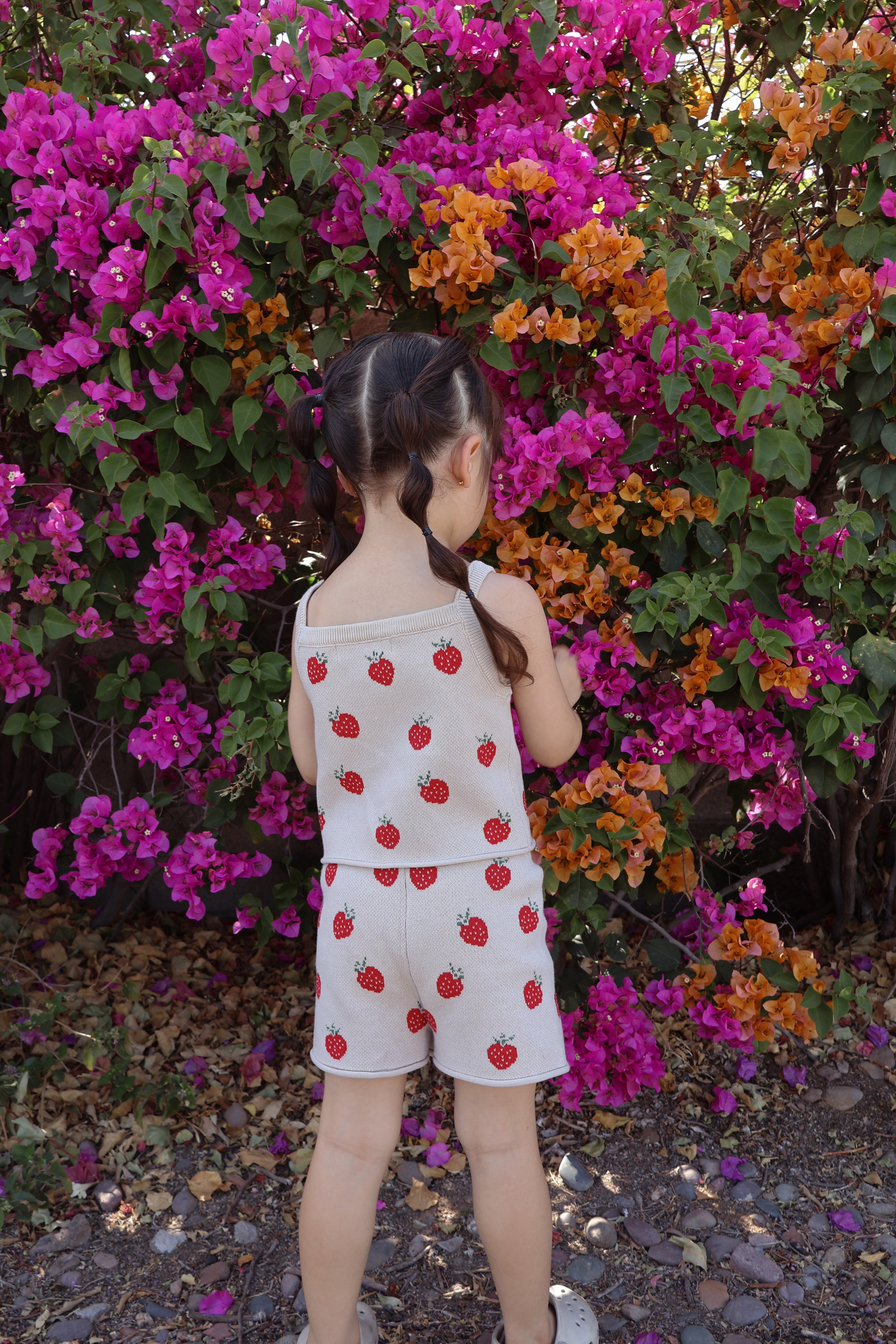 Girl wearing a strawberry knit two-piece set surrounded by colorful flowers, perfect for spring toddler clothes.